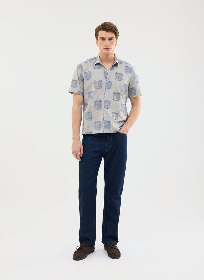 A man models the Percival Bruno Boxy Clerk Shirt in grey, featuring a patchwork design, styled with dark wash jeans.