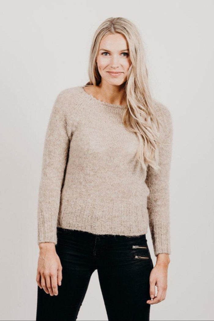 Woman wearing a beige sweater and black pants against a plain background