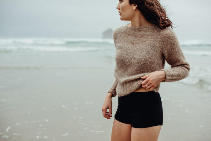 Woman wearing a beige sweater and black shorts standing on a beach.