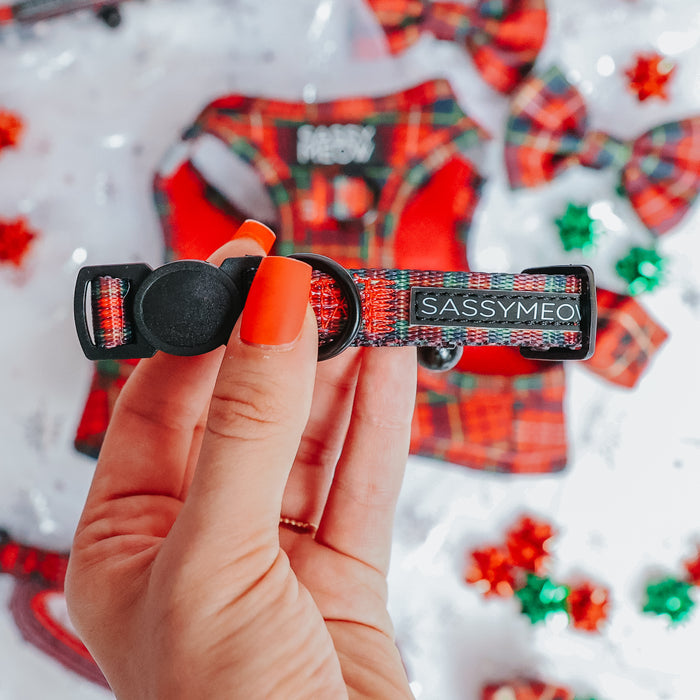 A hand with bright orange nail polish holds a Sassy Woof Cat Collar - Deck the Paws. Blurred in the background are matching holiday cat accessories and festive decorations, creating a cheerful scene.