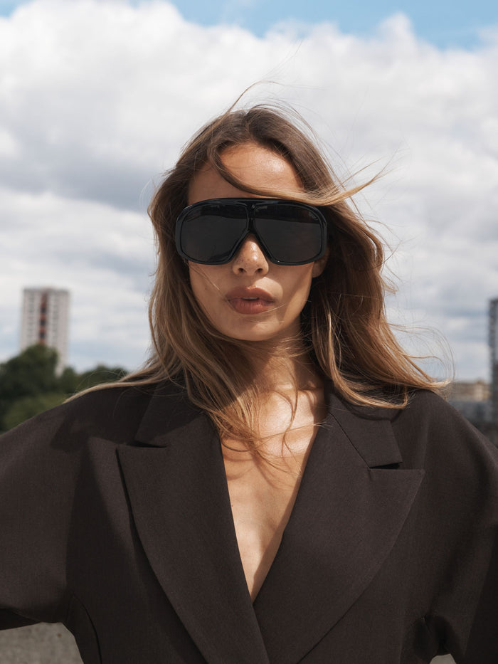 A woman outdoors wearing oversized black shield-style sunglasses and a dark brown blazer, with her hair blowing in the wind against a cloudy sky.