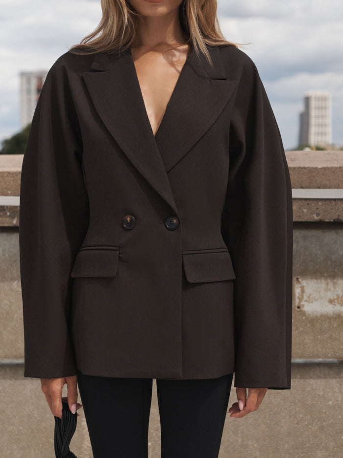 A woman wearing an oversized dark brown double-breasted blazer with notched lapels and flap pockets, paired with black pants, photographed outdoors against a blurred urban background.