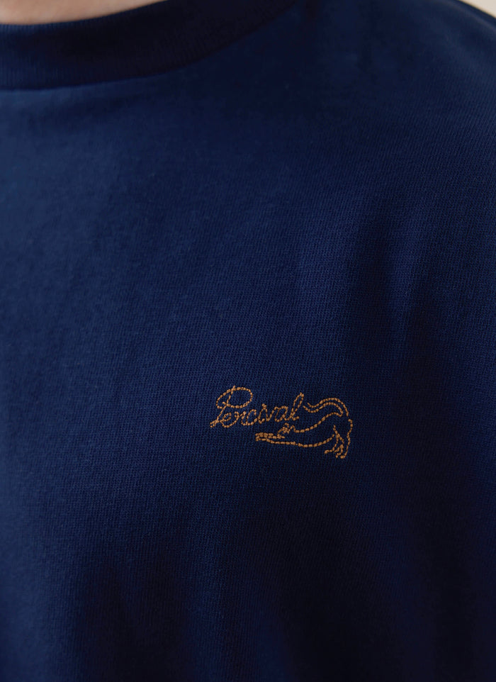 Gold-stitched Percival logo on the navy Clarke T-shirt, a piece from our organic cotton SS26 collection.