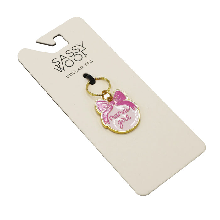 The Sassy Woof Collar Tag - Mama's Girl (Gold) is a round charm with a pink bow and "Mama’s Girl" in pink script, attached to a gold keyring and displayed on a cream brand card.