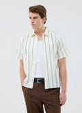A model wears the Percival Cross Stitch Boxy Clerk Shirt in ecru, styled open over a white vest with brown trousers.