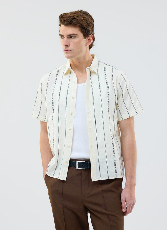 A model wears the Percival Cross Stitch Boxy Clerk Shirt in ecru, styled open over a white vest with brown trousers.