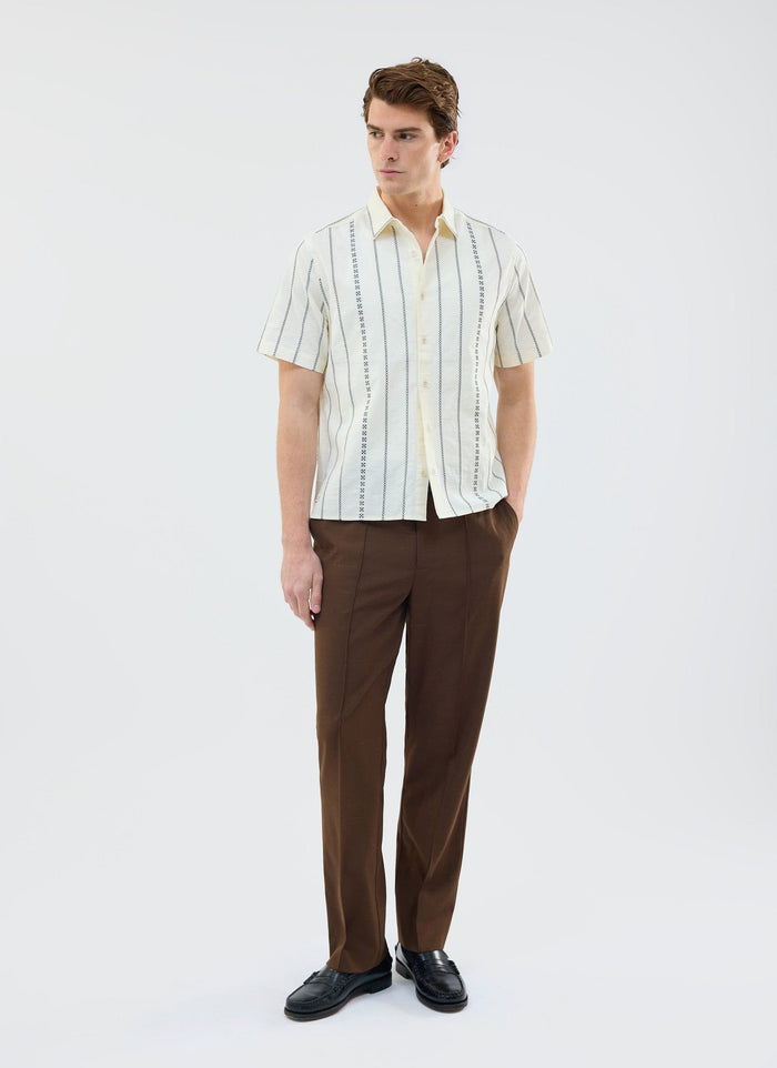A model wears the ecru Cross Stitch Boxy Clerk Shirt from Percival, styled with smart brown trousers and black loafers.