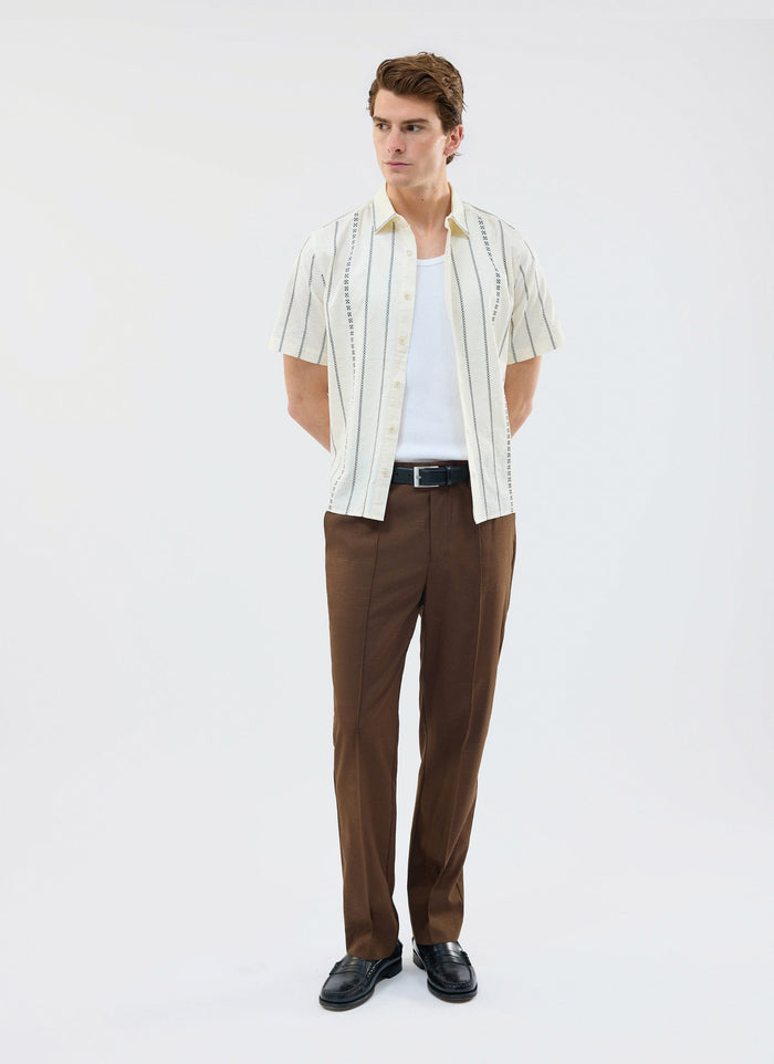 A man models the Percival Cross Stitch Boxy Clerk Shirt in ecru, worn open over a white vest with brown trousers.
