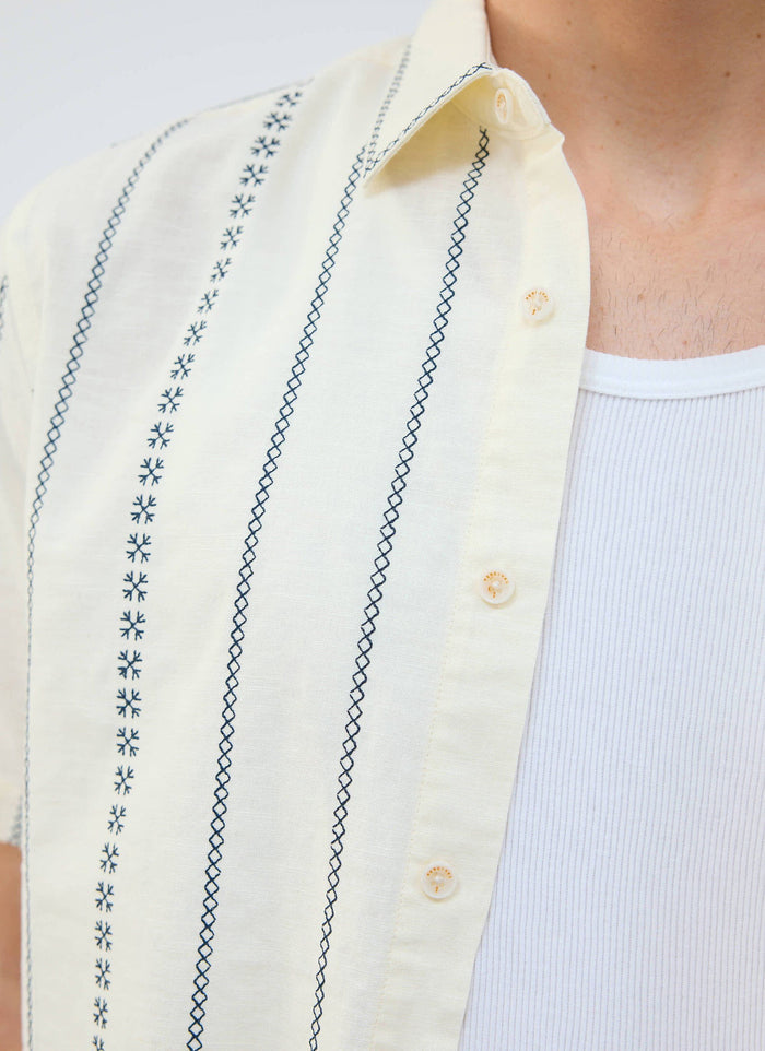 A close-up of the Percival Cross Stitch Boxy Clerk Shirt in ecru, showing the detailed blue embroidery and custom buttons.