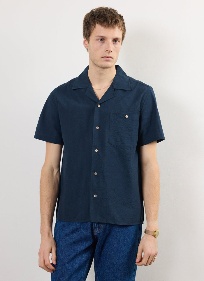 The Percival Cuban Shirt in navy seersucker, a textured short-sleeve shirt, shown on a model against a white wall.