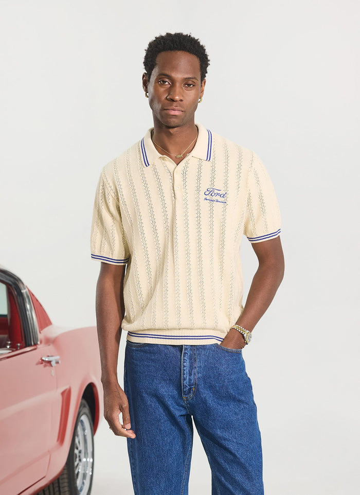 A model wears the Percival x Ford Dealership Knitted Polo in ecru with blue jeans, pictured in a studio with a red car.
