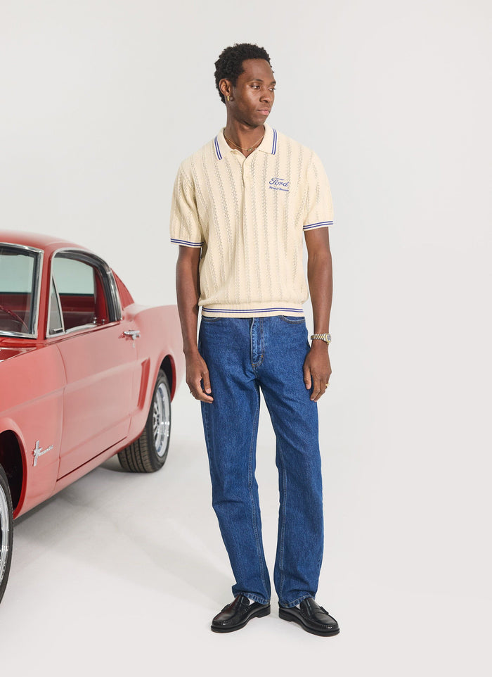 A model wears the ecru Dealership Knitted Polo from the Percival x Ford collaboration, next to a vintage red Ford car.