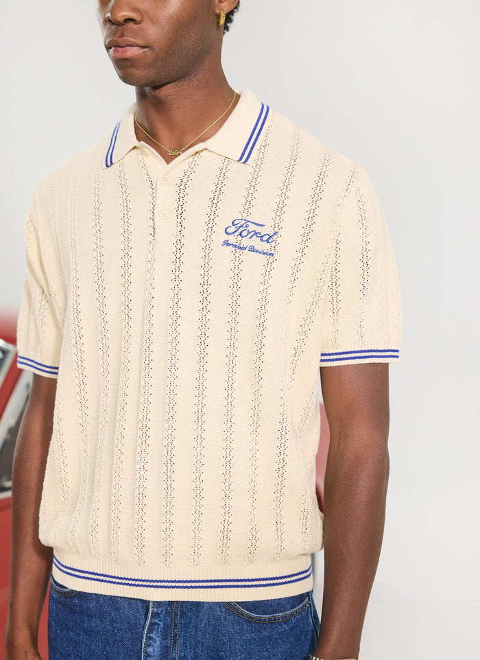 Close-up of the Percival x Ford Dealership knitted polo in ecru, showing the blue embroidered logo and striped tipping.