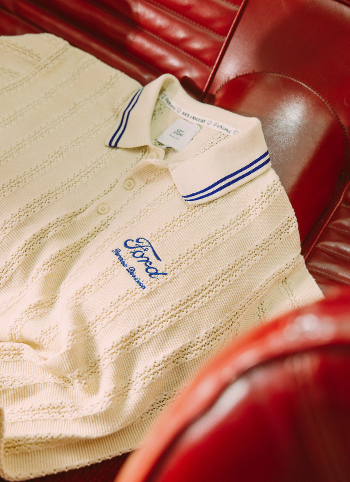 Close-up of the ecru Percival x Ford knitted polo, showing the blue Ford embroidery and collar detail, on a red car seat.