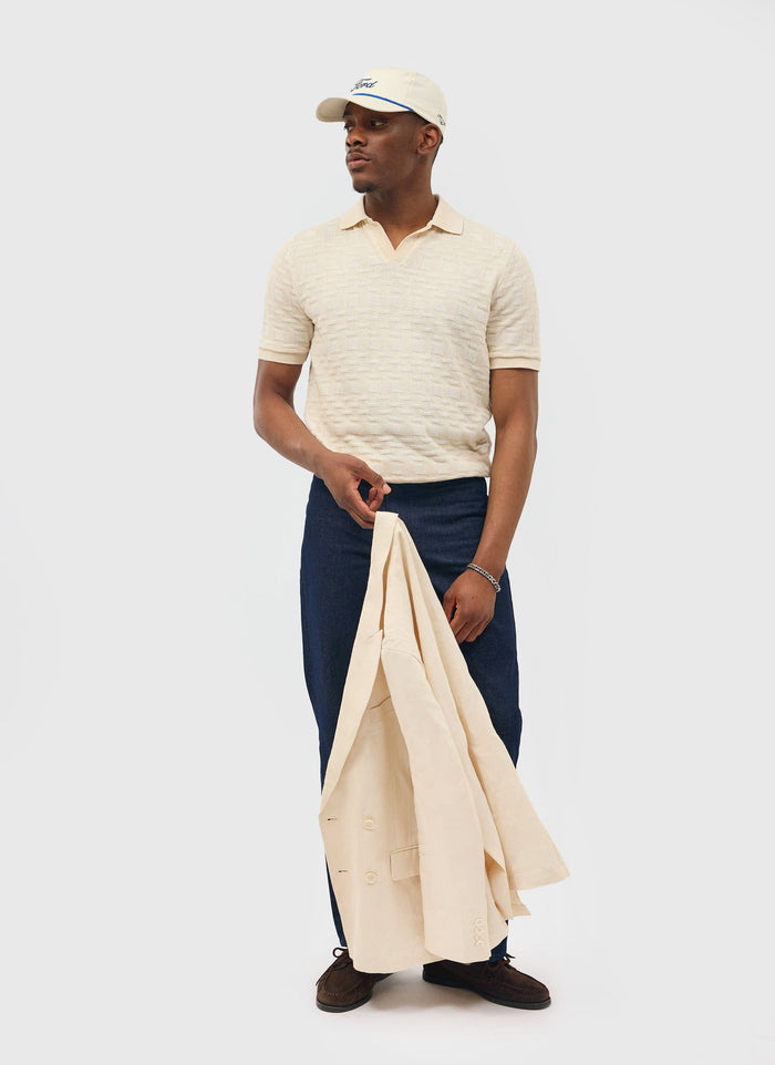 A model wears the Percival Deck Negroni Knitted Shirt in ecru with dark jeans, a cap, and holds a matching jacket.