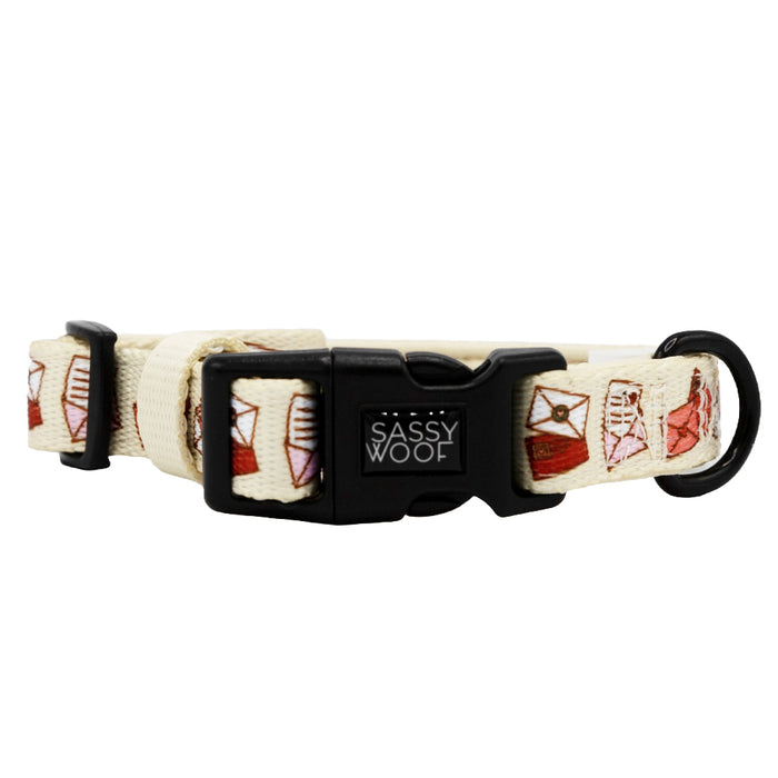 The Sassy Woof P.S. I Woof You dog collar is cream-colored with a small envelope pattern, an adjustable fit, black plastic buckle, and metal D-ring for leash attachment.