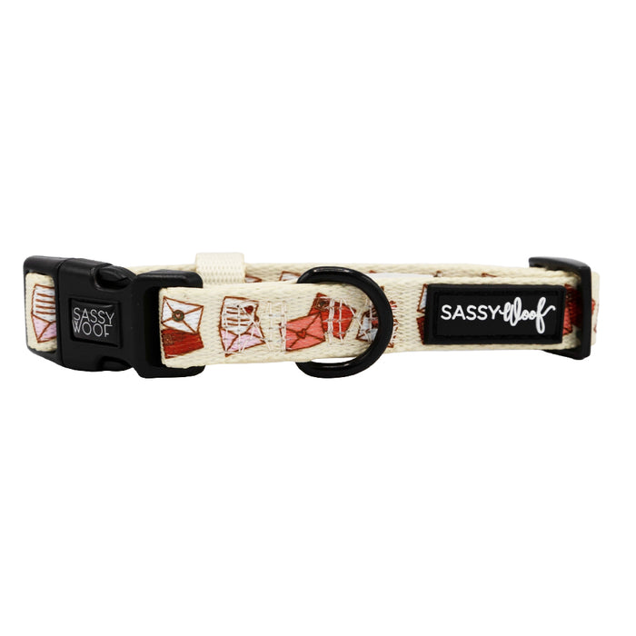 The Sassy Woof Dog Collar - P.S. I Woof You is a cream-colored adjustable collar with red and white dress pattern, black plastic buckle, metal D-ring, and a Sassy Woof logo tag—an adorable addition to your dog's accessories.