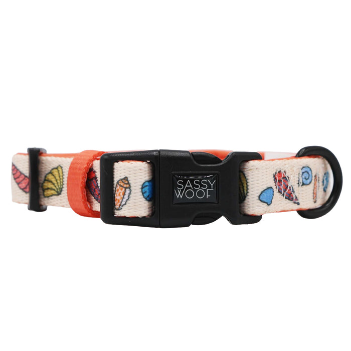 The Dog Collar - Shell Yeah by Sassy Woof features an adjustable cream strap with vibrant leaf and abstract patterns, orange accents, and a black buckle displaying the Sassy Woof brand name.