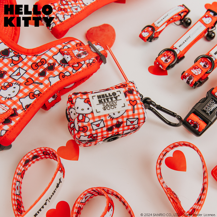 The Sassy Woof Dog Waste Bag Holder - Hello Kitty® Red Classic is shown with matching harness, leash, and bag dispenser featuring a red Hello Kitty pattern on a white surface with heart-shaped decor. HELLO KITTY appears in the top left corner.