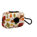 The Sassy Woof Dog Waste Bag Holder - Peanuts™ Harvest is a small rectangular accessory with fall-themed Snoopy, pumpkin, corn, and pie designs plus a black carabiner and round dispenser—ideal for stylish dog walks.