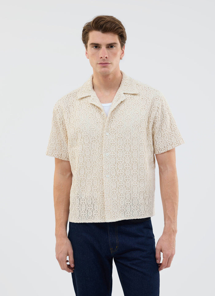 A man models the Percival Elio Crochet Cuban Shirt in ecru, styled over a white vest and paired with dark wash jeans.