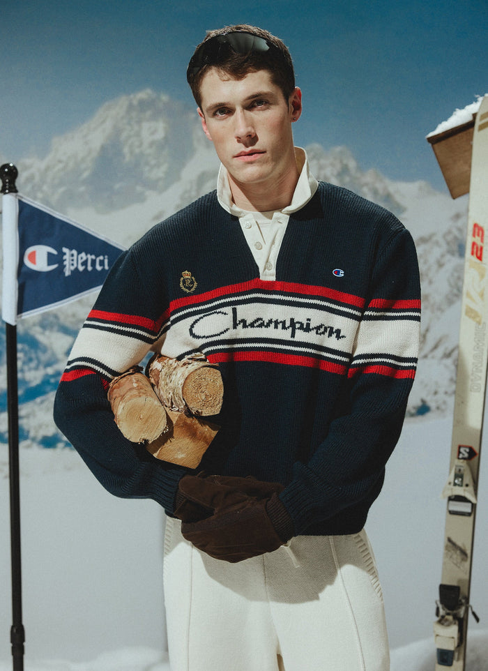 A model wears the navy Champion x Percival Jacquard Knitted Rugby Polo Shirt, holding logs in a snowy ski resort setting.