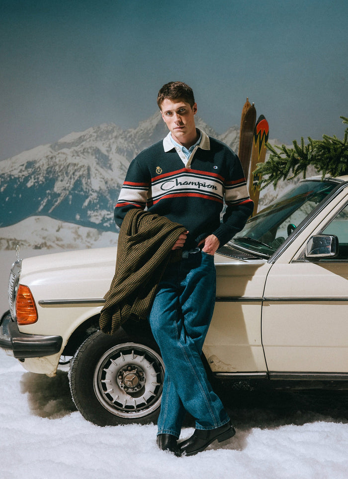 A man models the Champion x Percival navy knitted rugby shirt, leaning against a vintage car in a snowy, mountain scene.