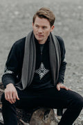 Man sitting on a log wearing a dark sweater with a patterned scarf and black pants.