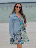 Woman in a floral dress and denim jacket standing on a beach.