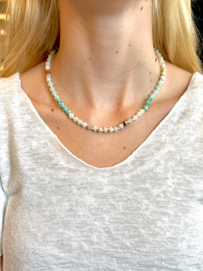 Beaded necklace with faceted aqua, jade, and pearl-toned beads and a gold extender chain, styled on a woman wearing a white top.