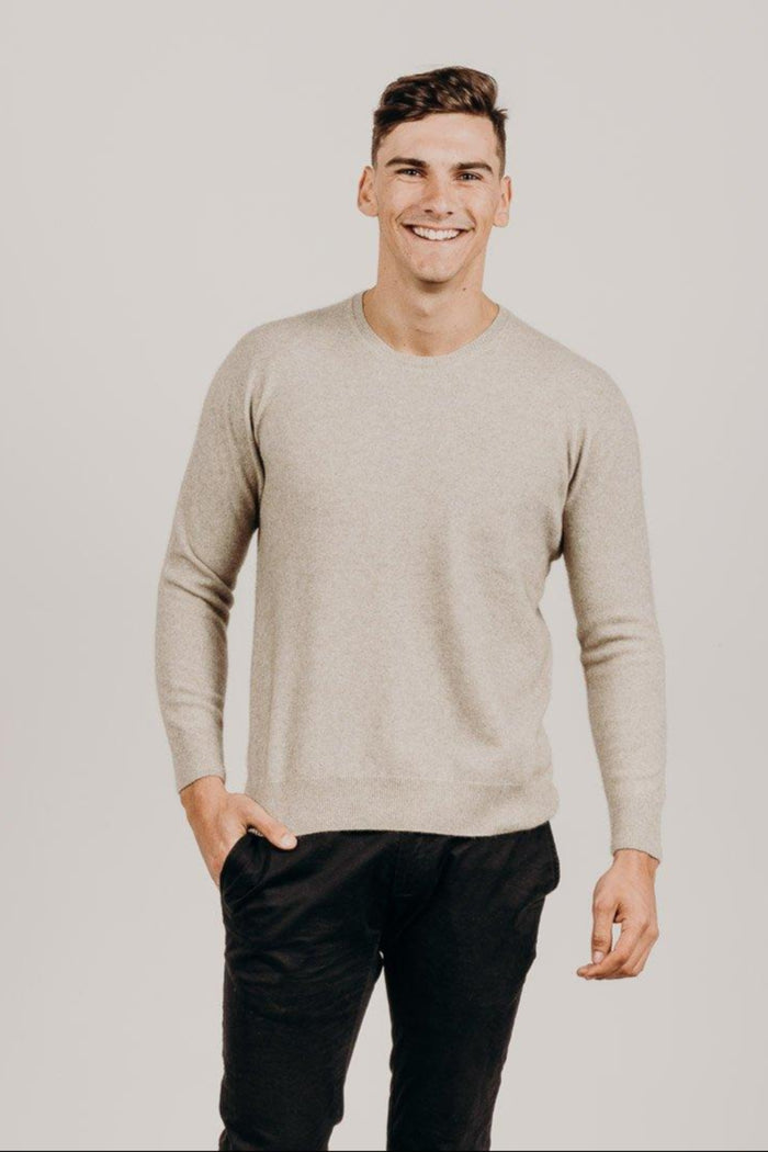 Man wearing a beige sweater and black pants against a plain background