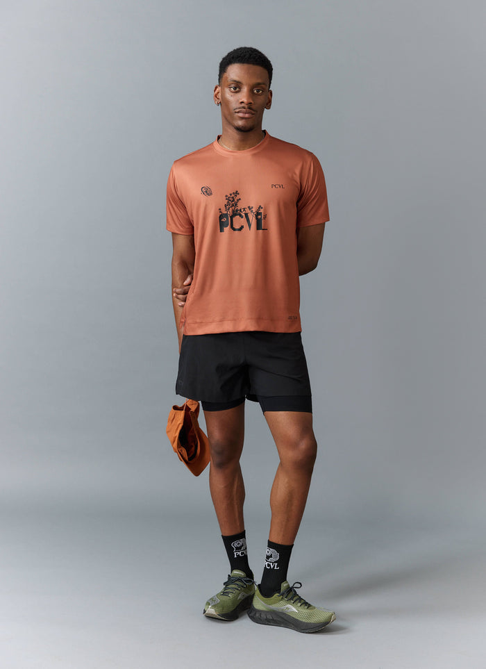 A model wears the rust PCVL Printed Performance Top from Percival, styled with black shorts, socks, and a matching cap.