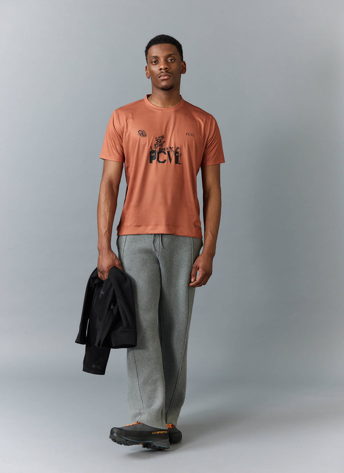A model wears the Percival PCVL Printed Performance Top in rust, styled with grey trousers and holding a black jacket.