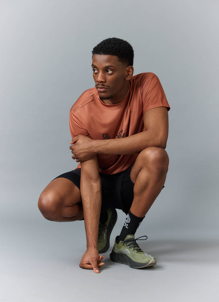 A model squats in a studio wearing the rust PCVL Printed Performance Top from Percival, with black shorts and trainers.