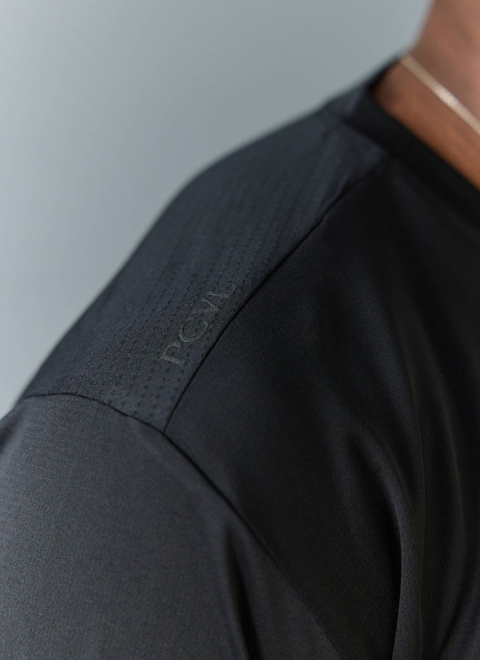 A close-up of the tonal 'PCVL' branding on the shoulder of the black Long Sleeve Printed Performance Top by Percival.
