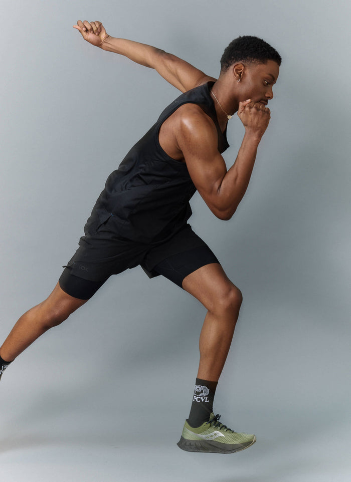 A model in a running pose wears the black PCVL Performance Vest and matching athletic shorts from Percival.