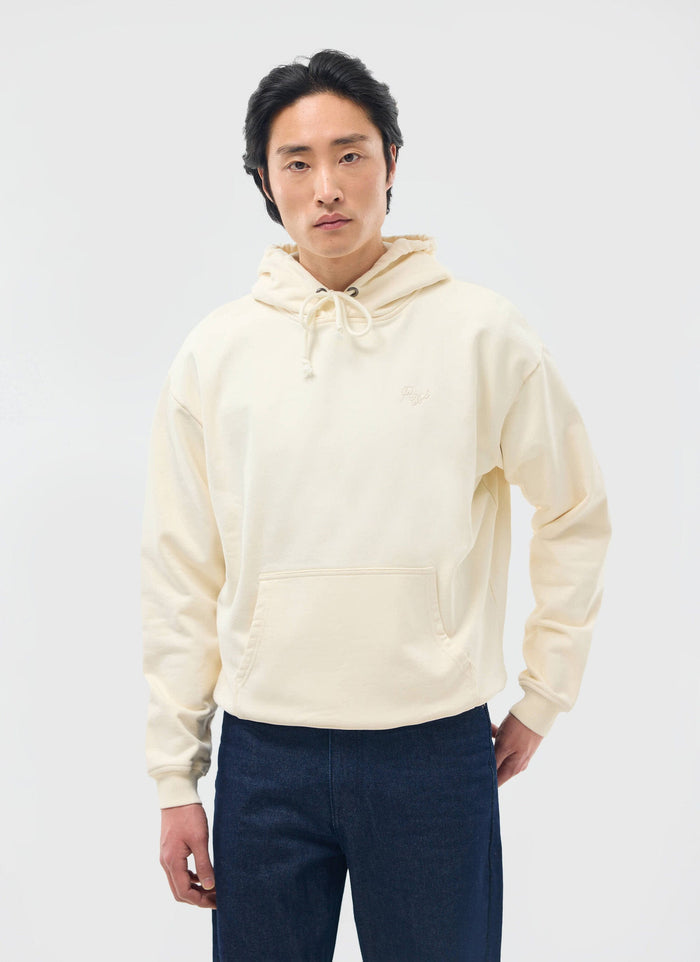 A man models the Percival Perci Chainstitch Hoodie in ecru, showcasing its relaxed fit with dark denim jeans.