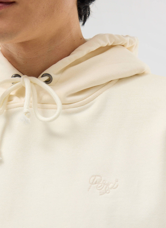 A close-up of a model wearing the Percival Perci Chainstitch Hoodie in ecru, showing the tonal chest embroidery.