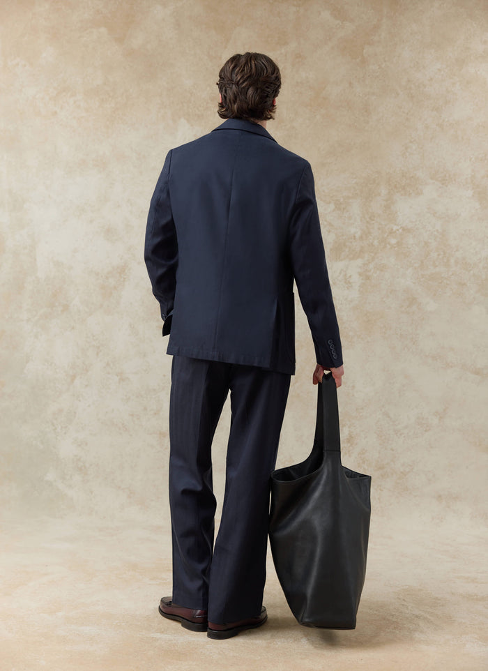 Rear view of a model wearing navy Percival lyocell pleated trousers and matching blazer, holding a large black bag.
