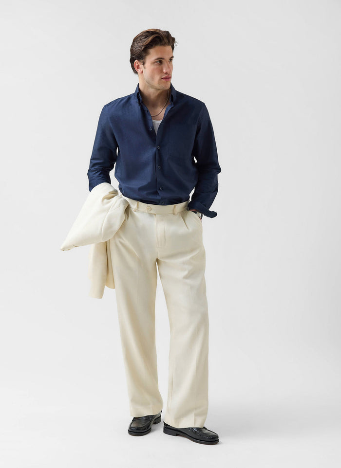 A man models the Percival Pleated Tailored Trousers in cream, styled with a navy shirt and holding the matching blazer.