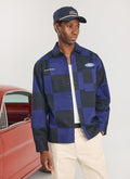 A model wears the Percival x Ford navy and black checkerboard overshirt with cream trousers and a navy cap.