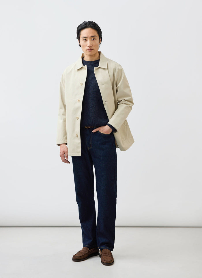A man models the Percival Sherlock Trench Coat in stone, styled with a dark blue knit, jeans, and loafers.
