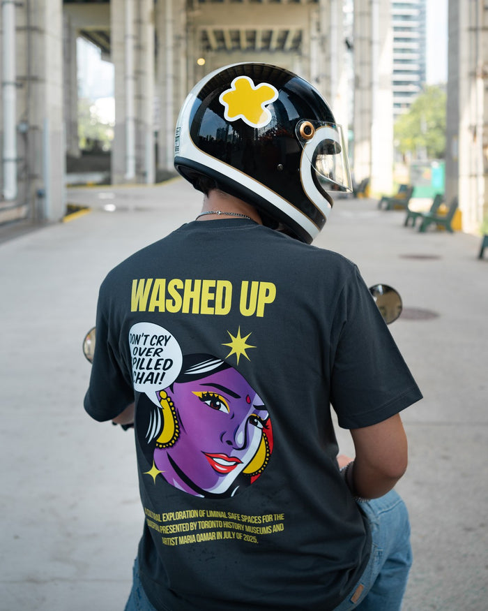Person wearing a gray t-shirt with colorful graphics and text, sitting on a motorcycle.