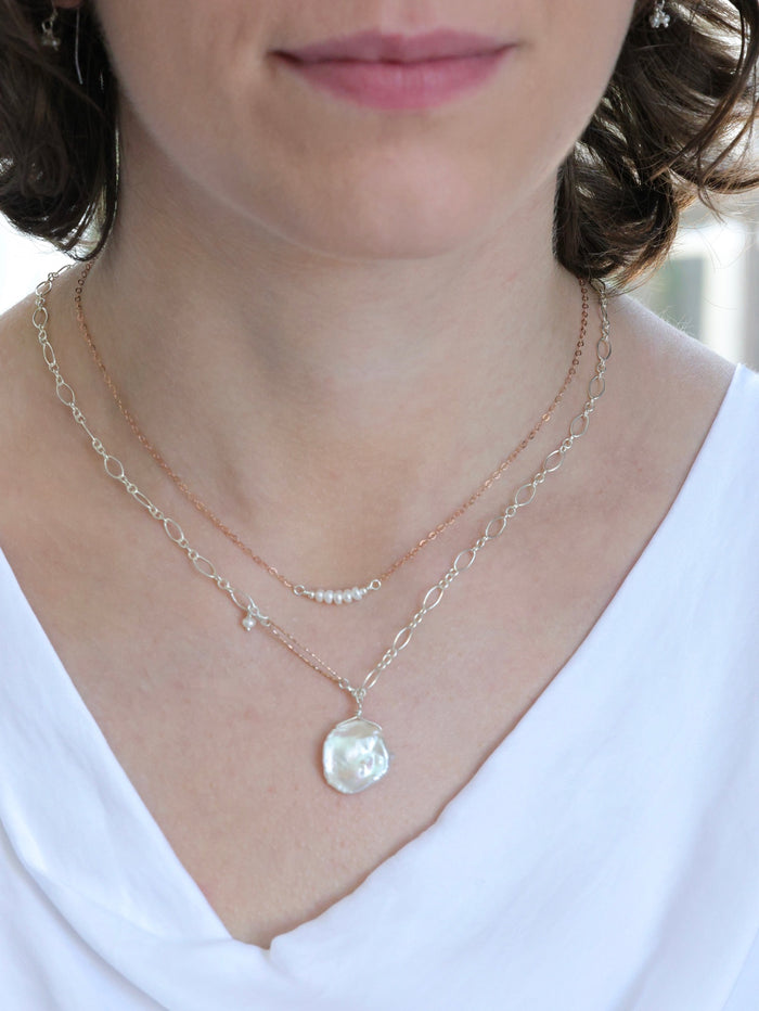 Close-up of a woman wearing layered necklaces from Ardor Boutique—one with five dainty pearls on a rose gold chain, and a second with a large coin pearl pendant on a silver chain—styled with a white blouse for a refined, elegant look.