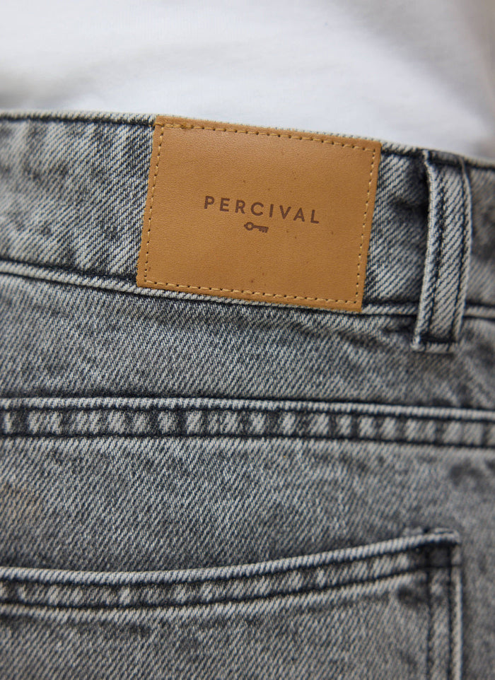 A close-up of the Percival Wide Leg Denim Jeans in warm grey wash, focusing on the tan leather logo patch.